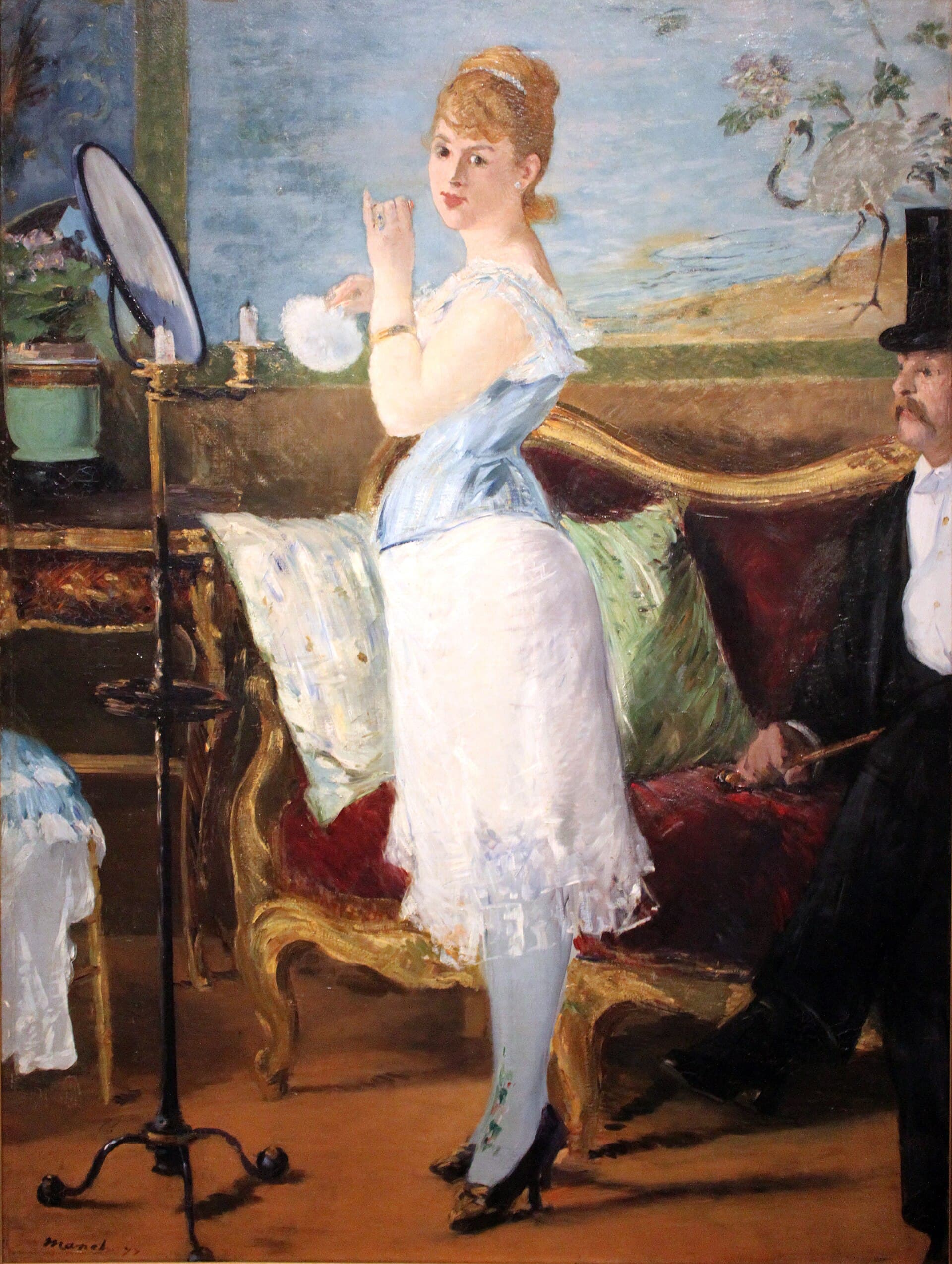 A young woman in a corset and petticoat at her mirror, powder puff in hand, a seated gentleman cropped at the right.